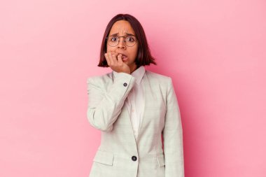 Pembe arka planda izole edilmiş, tırnak yiyen, gergin ve endişeli yeşil takım elbiseli genç bir ırk kadını..