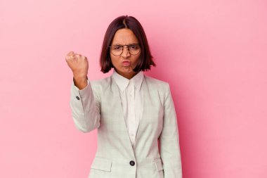 Pembe arka planda izole edilmiş yeşil takım elbiseli genç bir ırk kadını kameraya yumruk gösteriyor, agresif bir yüz ifadesi..