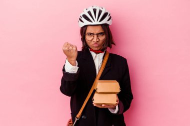 Genç iş kadını, bisiklet kaskı takıyor, elinde hamburgerle pembe arka planda izole edilmiş, kameraya yumruk, agresif yüz ifadesi gösteriyor..