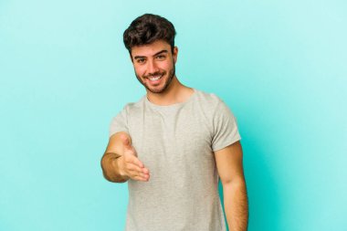 Mavi arka planda izole edilmiş genç beyaz adam selamlama jesti olarak kameraya elini uzatıyor..