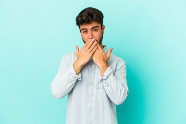 Mavi arka planda izole edilmiş genç beyaz adam elleriyle ağzını kapatırken şok oldu..