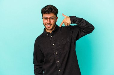 Mavi arka planda izole edilmiş genç beyaz adam bir gömlek fotokopi alanını işaret ediyor, gururlu ve kendinden emin.