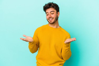 Kafkasyalı genç adam mavi arka planda izole edilmiş. Elinde avuç içi olan bir şey var ve kameraya sunuyor..