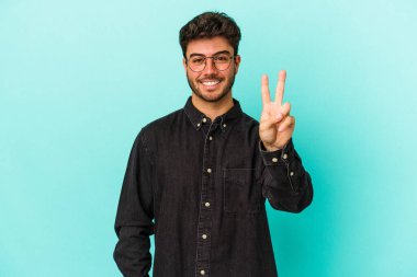 Mavi arka planda izole edilmiş genç beyaz adam zafer işareti gösteriyor ve gülümsüyor..