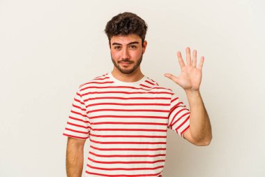 Beyaz arka planda izole edilmiş genç beyaz bir adam gülümseyerek beş numarayı parmaklarıyla gösteriyor..