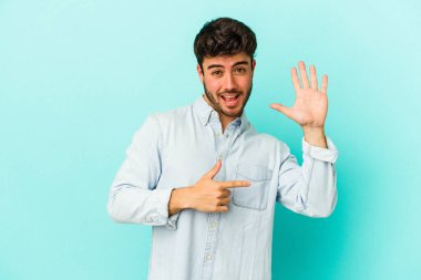 Mavi arka planda izole edilmiş genç beyaz adam gülümseyerek beş numarayı parmaklarıyla gösteriyor..