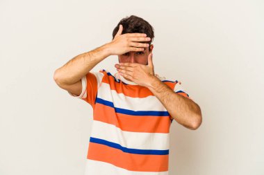 Beyaz arka planda izole edilmiş genç beyaz bir adam kameraya parmaklarıyla göz kırpıyor, yüzünü kapatmaktan utanıyor..