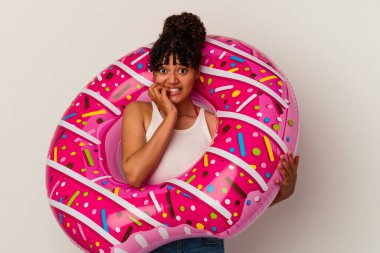 Beyaz arka planda izole edilmiş, tırnak yiyen, gergin ve kaygılı şişirilebilir bir donut tutan genç bir ırk kadını..