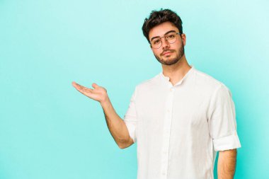 Mavi arka planda izole edilmiş genç beyaz bir adam bir palmiye üzerinde fotokopi alanı ve belinde bir el daha gösteriyor..