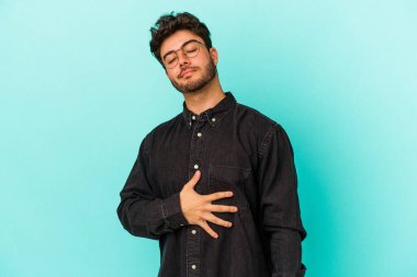 Mavi arka planda izole edilmiş genç beyaz adam karnına dokunuyor, nazikçe gülümsüyor, yemek yiyor ve tatmin oluyor..