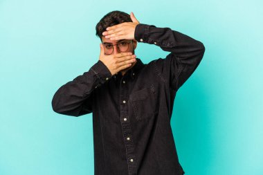 Mavi arka planda izole edilmiş genç beyaz adam kameraya parmaklarıyla göz kırpıyor, yüzünü kapatmaktan utanıyor..