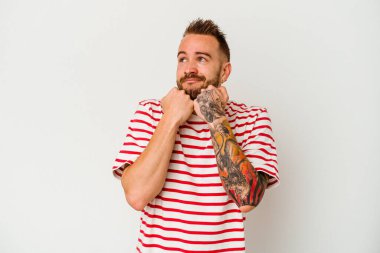 Beyaz arka planda izole edilmiş genç dövmeli beyaz adam ellerini çenesinin altında tutuyor ve mutlu bir şekilde yan tarafa bakıyor..