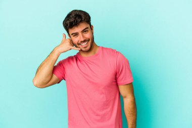 Mavi arka planda izole edilmiş genç beyaz adam parmaklarıyla bir cep telefonu hareketi gösteriyor..