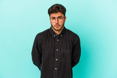 Mavi arka planda izole edilmiş genç beyaz adam omuzlarını silkeliyor ve gözleri açık..