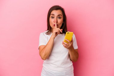 Pembe arka planda cep telefonu tutan orta yaşlı beyaz kadın bir sır saklıyor ya da sessizlik istiyor..