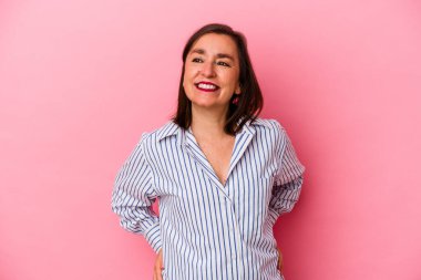 Orta yaşlı, beyaz bir kadın pembe arka planda izole edilmiş. Kendine güveni tam. Ellerini kalçasında tutuyor..