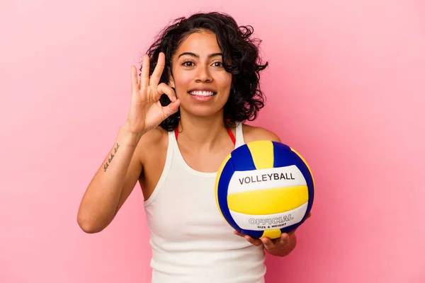 Young latin woman holding a volley ball isolated on pink background cheerful and confident showing ok gesture.