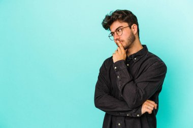 Mavi arka planda izole edilmiş genç beyaz adam şüpheli ve şüpheci bir ifadeyle yana bakıyor..