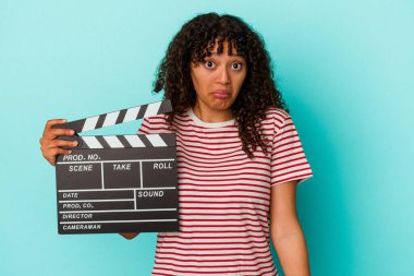 Young mixed race woman holding a clapperboard isolated on blue background shrugs shoulders and open eyes confused. clipart