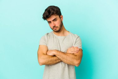 Kafkasyalı genç adam mavi arka planda izole edilmiş hoşnutsuzca kaşlarını çatmış, kollarını kavuşturmuş..