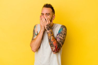 Sarı arka planda izole edilmiş genç dövmeli beyaz adam bir şeye gülüyor, elleri ağızda..