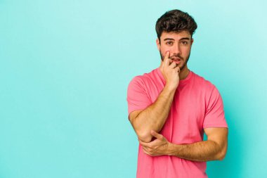 Mavi arka planda izole edilmiş genç beyaz adam şüpheli ve şüpheci bir ifadeyle yana bakıyor..