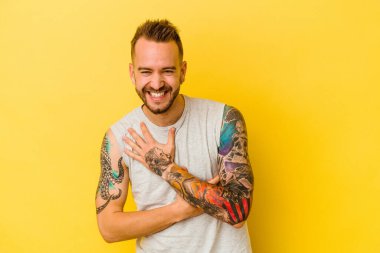 Sarı arka planda izole edilmiş genç dövmeli beyaz adam mutlu bir şekilde gülüyor ve ellerini karnının üstünde tutarak eğleniyor..