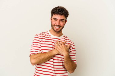 Beyaz arka planda soyutlanmış beyaz tenli genç bir adam gülüyor, elini yüreğine koyuyor, mutluluk kavramını koruyor..