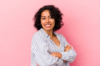 Pembe arka planda soyutlanmış genç kıvırcık Latin kadın. Kendine güveniyor, kararlılıkla kollarını kavuşturuyor..