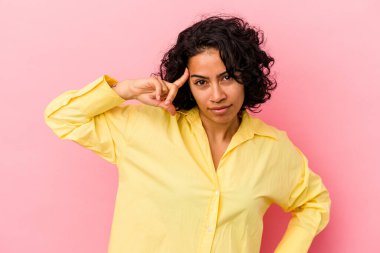 Pembe arka planda izole edilmiş genç kıvırcık Latin kadın parmağıyla tapınağı işaret ediyor, düşünüyor ve bir göreve odaklanıyor..