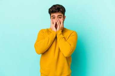 Mavi arka planda izole edilmiş genç beyaz adam şok olmuş, ağzı ellerle kaplanmış, yeni bir şey keşfetmeye hevesli..