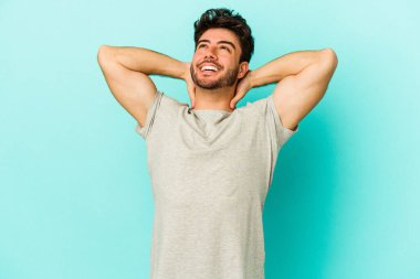 Kafkasyalı genç adam mavi arka planda izole edilmiş. Kendine güveniyor, elleri başının arkasında..