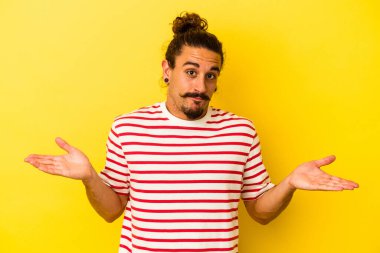 Sarı arka planda izole edilmiş, uzun saçlı, genç, beyaz bir adam. Kafası karışmış ve omuz silkiyor..