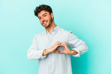Mavi arka planda izole edilmiş genç beyaz adam gülümsüyor ve elleriyle kalp şeklini gösteriyor..