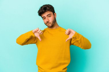 Mavi arka planda izole genç beyaz adam baş parmağını aşağı gösteriyor, hayal kırıklığı konsepti.
