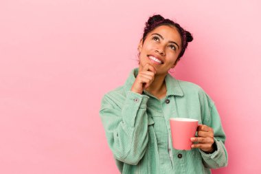 Pembe arka planda bir kupa tutan genç Latin kadın şüpheli ve şüpheci bir ifadeyle yana bakıyor..