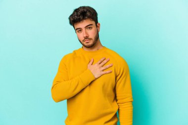 Kafkasyalı genç adam mavi arka planda izole edilmiş yemin ediyor, elini göğsüne koyuyor..