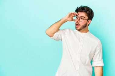 Kafkasyalı genç adam mavi arka planda izole edilmiş, uzaklara bakıyor ve elini alnına koyuyor..