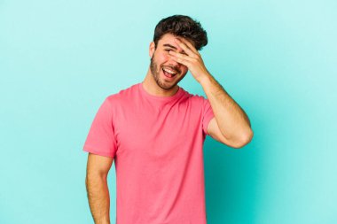 Mavi arka planda soyutlanmış genç beyaz bir adam elleriyle gözlerini kapatıyor, geniş bir gülümsemeyle bir sürpriz bekliyor..