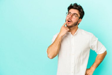 Mavi arka planda izole edilmiş genç beyaz adam şüpheli ve şüpheci bir ifadeyle yana bakıyor..