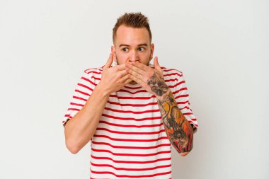 Beyaz arka planda izole edilmiş genç dövmeli beyaz adam elleri ile ağzını kapatırken şok oldu..