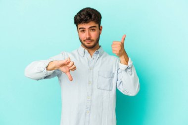 Kafkasyalı genç adam mavi arka planda izole edilmiş, baş parmağını kaldırıyor, zor seçim konsepti.