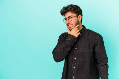 Kafkasyalı genç adam mavi arka planda izole edilmiş kafasının arkasına dokunuyor, düşünüyor ve bir seçim yapıyor..