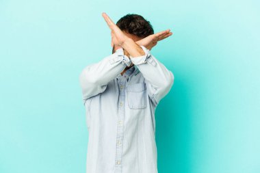 Kafkasyalı genç adam mavi arka planda izole edilmiş iki kolu çapraz tutuyor, inkar kavramı.