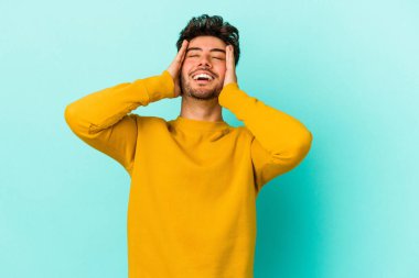 Mavi arka planda izole edilmiş genç beyaz adam ellerini başının üstünde tutarak neşe içinde gülüyor. Mutluluk kavramı.