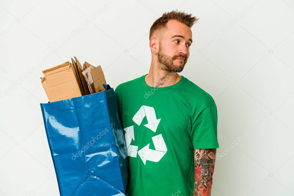 Young caucasian tattooed man recycled cardboard isolated on white background looks aside smiling, cheerful and pleasant.