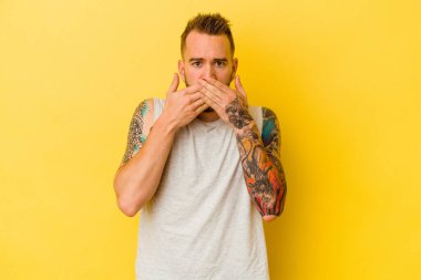Sarı arka planda izole edilmiş genç dövmeli beyaz adam elleri ile ağzını kapatırken şok oldu..