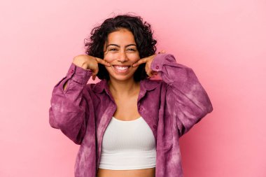 Pembe arka planda izole edilmiş genç kıvırcık Latin kadın gülümsüyor, parmaklarını ağzına doğrultuyor..