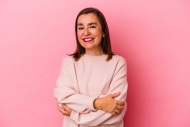 Pembe arka planda soyutlanmış orta yaşlı beyaz bir kadın. Kendine güveniyor, kararlılıkla kollarını kavuşturuyor..