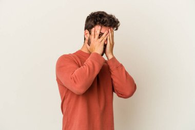 Beyaz arka planda izole edilmiş genç beyaz bir adam parmaklarının arasından korkmuş ve gergin bir şekilde göz kırpıyor..